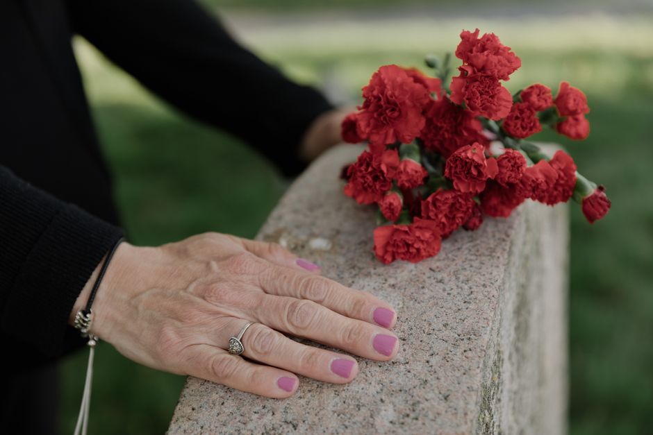 A person's hand with pink-painted nails resting on a stone ledge, beside a small bouquet of vibrant red carnations with ruffled petals. The bouquet is wrapped in clear plastic, and the person's hand i
