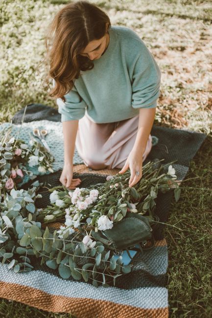 A woman with long, wavy brown hair dressed in a light blue sweater and beige skirt is kneeling on a dark woven blanket outdoors, arranging a floral bouquet. The bouquet features soft pink and white ro