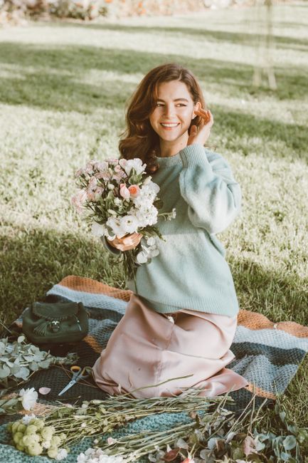 A young woman with long, wavy brown hair and a bright smile is sitting on a multicolored blanket spread on a lush green lawn in the park. She is holding a bouquet of fresh flowers that includes white 
