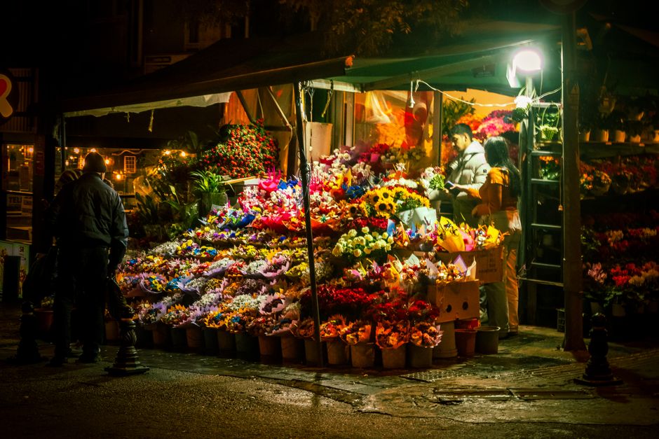 At a flower stall illuminated by warm lighting at night, a variety of fresh flowers including sunflowers, roses, lilies, and carnations in vibrant shades of yellow, pink, red, and white are arranged d