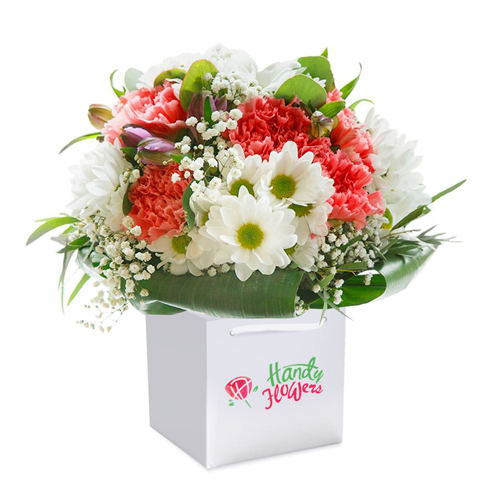 Presented in a contemporary white square gift bag, this vibrant bouquet brings together the distinct character and artistry of Welwyn's flower shops. The bouquet brims with lush coral, peach, and blazing red carnations, their abundant ruffled petals gathered in a rounded, generous shape that feels both festive and inviting. Interspersed among these are immaculate white daisies, each radiant with a yellow-green center, their clean beauty recalling meadows near Stanborough Park. Wisps of white gypsophila add a touch of softness, their delicate blossoms floating between the main flowers, while a handful of purple-dusted, unopened buds introduce drama and visual depth on the bouquet's left. Deep green, broad-leaved foliage encircles the arrangement, forming a polished collar that accentuates the bouquet's vibrancy. The white bag's ribbon handles offer an elegant finishing touch, suggesting easy transport through Welwyn Garden City's lanes. Brightly lit, every tonal nuance and surface is revealed: petals gleam softly, foliage shines, and the composition radiates freshness, making it a memorable gift for birthdays, anniversaries, or as a thoughtful surprise across Welwyn's neighborhoods. This hand-crafted floral gift showcases expert design and the welcoming nature of Welwyn's florists.